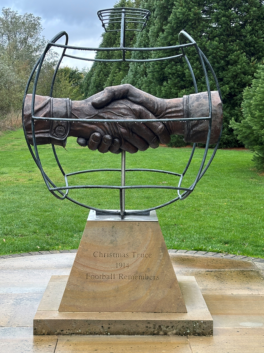 The Christmas Truce Memorial, bearing on it's base, the words, 'Christmas Truce 1914 Football Remembers'. Sitting atop the base, is a metal fromework depicting a footballwith two hands comming together in its centre in a handshake.